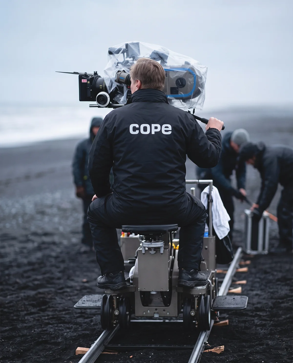 Camera operator in the cope windbreaker on a dolly at a black-sand beach shoot