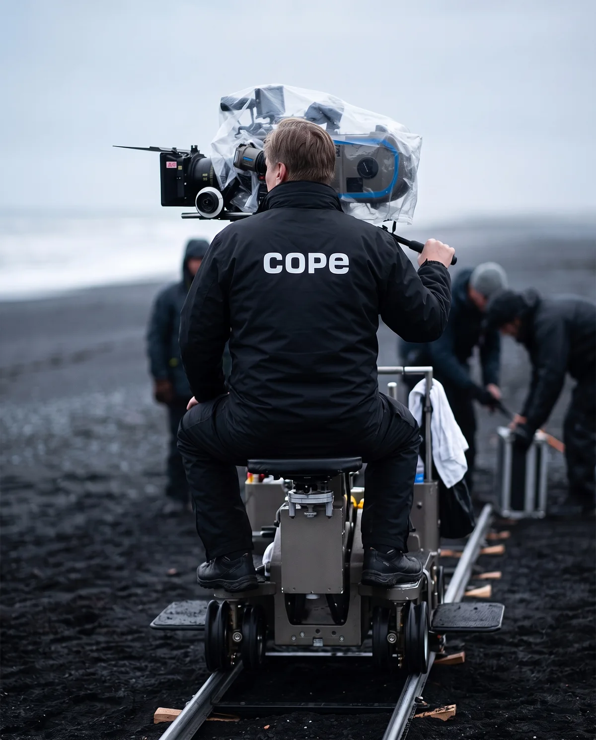 Camera operator in the cope windbreaker on a dolly at a black-sand beach shoot